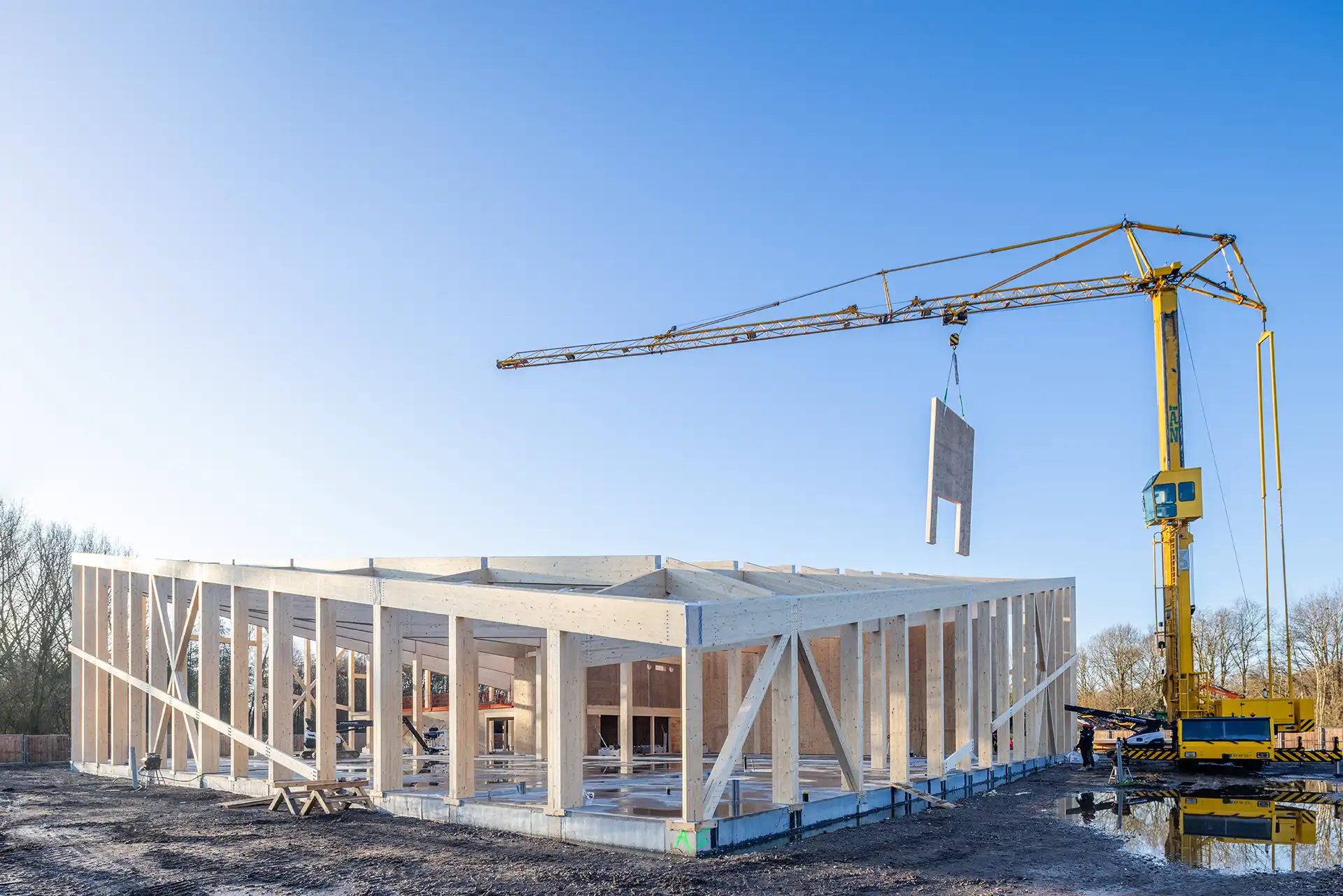 Een gele bouwkraan hijst een betonnen wanddeel bij een houten skelet van een gebouw onder een heldere blauwe hemel.
