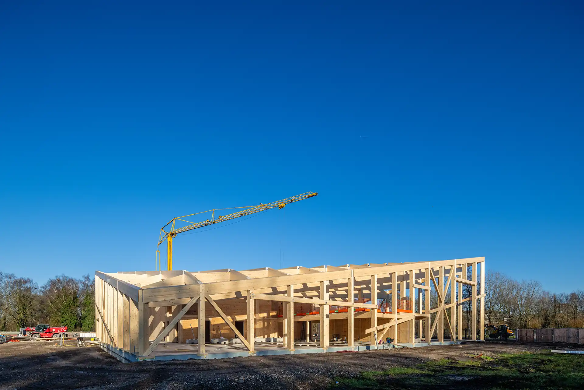 Houten skelet van een gebouw in aanbouw op een bouwplaats met een gele kraan en een heldere blauwe lucht.