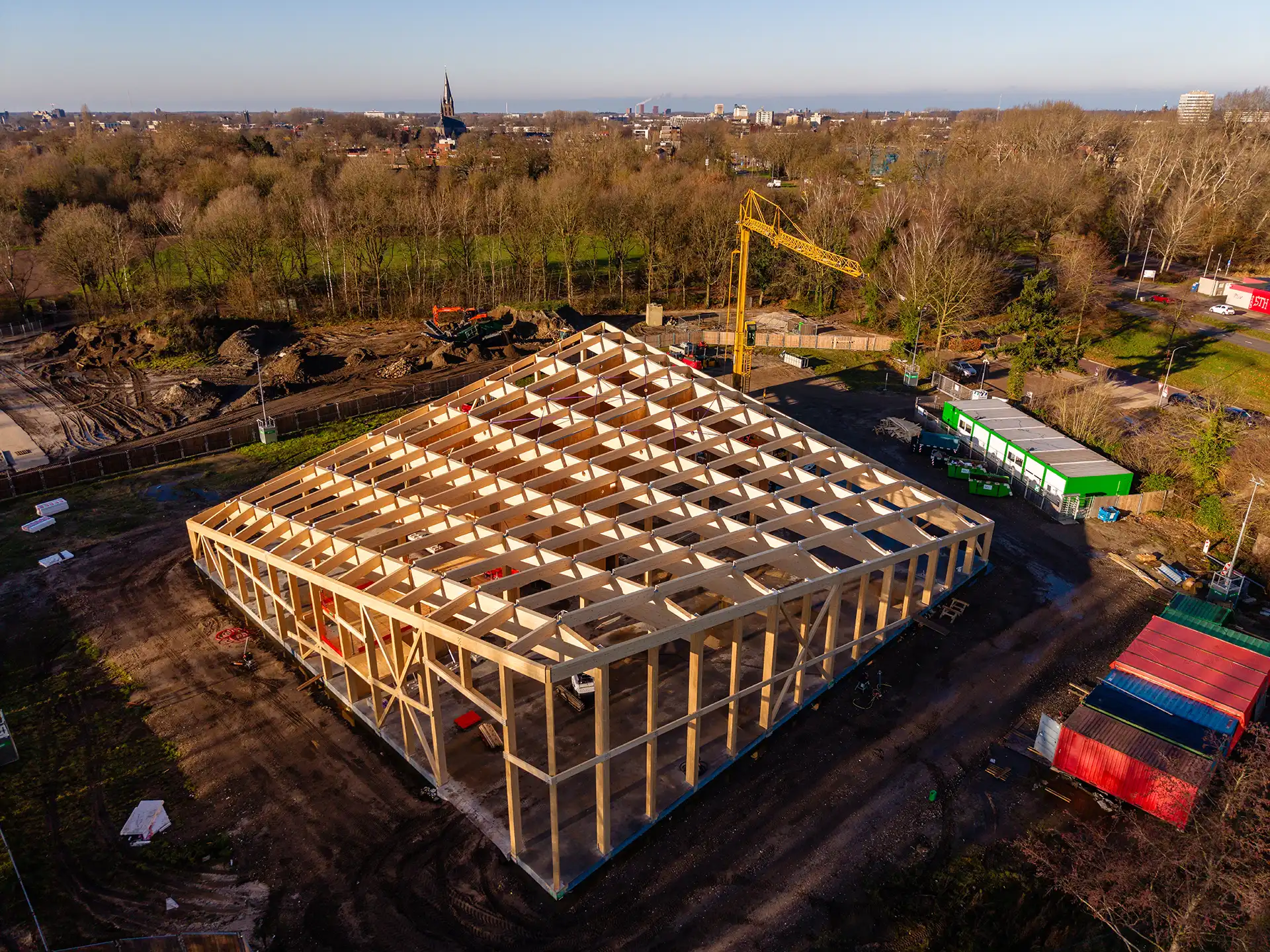 Luchtfoto van de houten structuur van een gebouw in aanbouw op een bouwplaats met een hijskraan en groene containers.