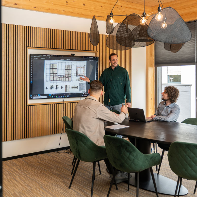 Drie mannen bespreken een bouwplan op een scherm in een moderne vergaderruimte met houten accenten en groene stoelen.