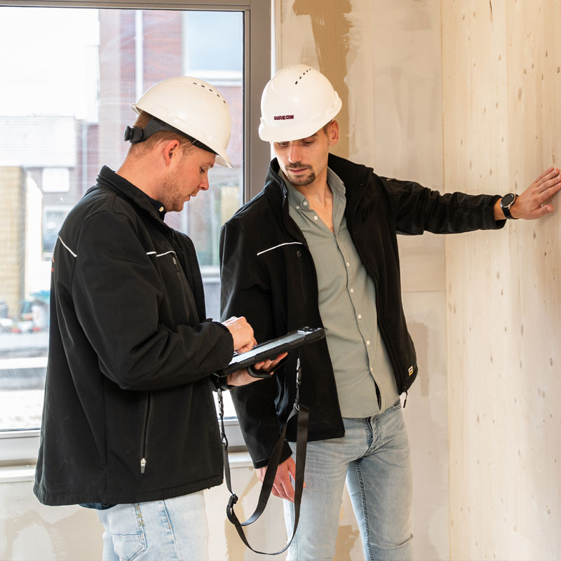 Twee bouwvakkers met witte helmen en zwarte jassen bekijken een tablet in een ongefinisseerde kamer.