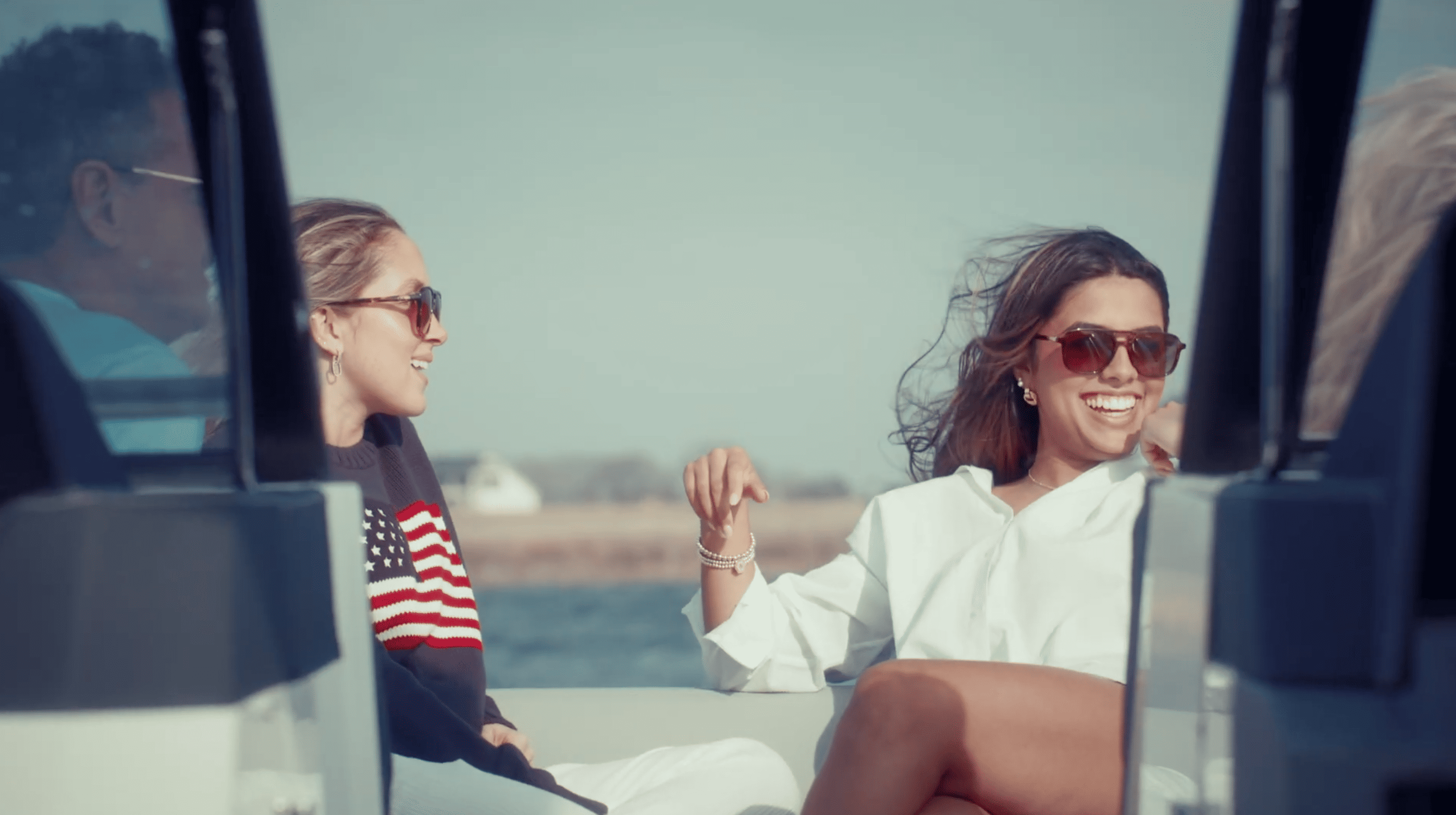 Two women wearing sunglasses smiling and chatting while sitting on a boat with water and a shoreline in the background.
