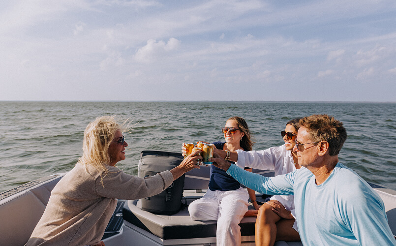 Four adults wearing sunglasses toasting canned drinks while sitting on a boat with ocean background under a partly cloudy sky.