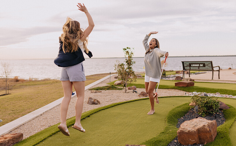 Two women happily celebrating on a mini golf course near a water body at sunset.