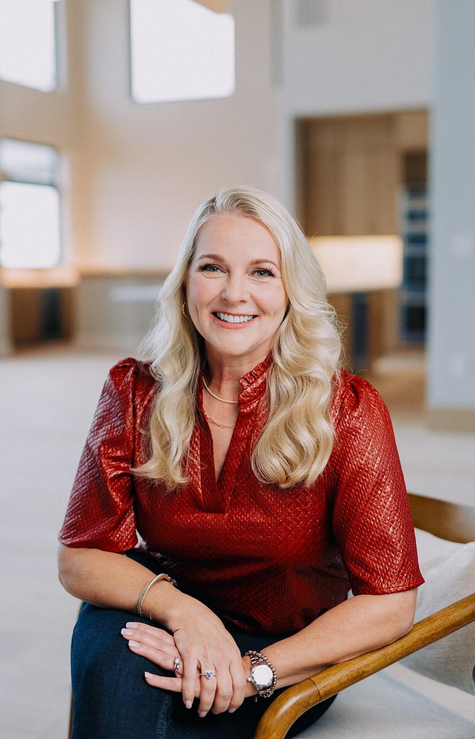 Smiling woman with long blonde hair wearing a textured red blouse and sitting on a wooden chair indoors.