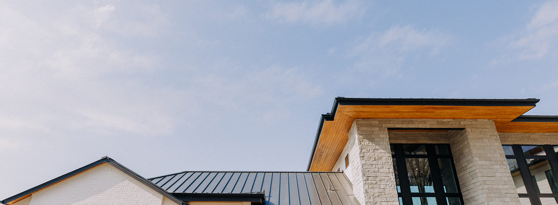 Modern house roof with light stone walls and large black-framed windows under a partly cloudy blue sky.