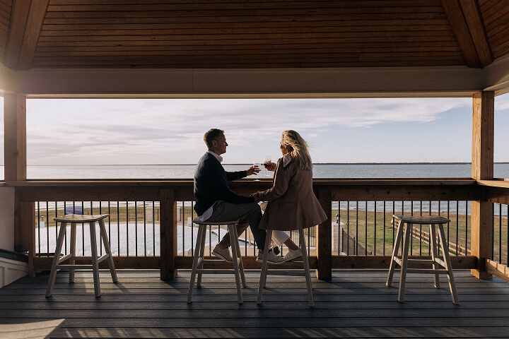 Couple sitting on stools under a wooden gazebo, holding wine glasses and facing a waterfront view at sunset.