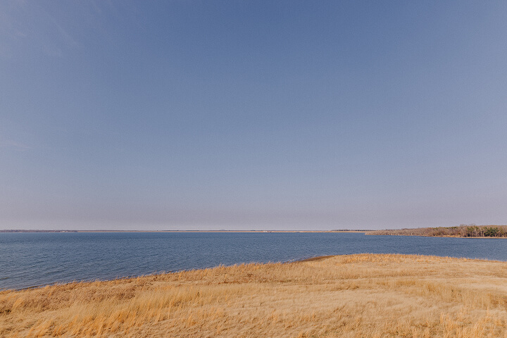 Wide view of a calm blue lake with golden grassy shore under a clear blue sky.