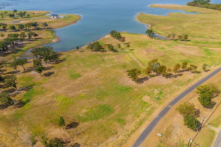 Aerial view of a large grassy landscape with scattered trees, a winding paved road, and a body of water in the background.