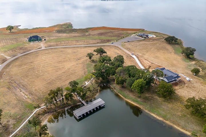 Aerial view of a lakeside area with dry grassy fields, scattered trees, a covered dock extending into the water, and a few buildings near the shore.