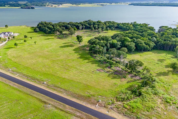 Aerial view of a green grassy landscape with scattered trees, a small cluster of houses, and a large body of water in the background.