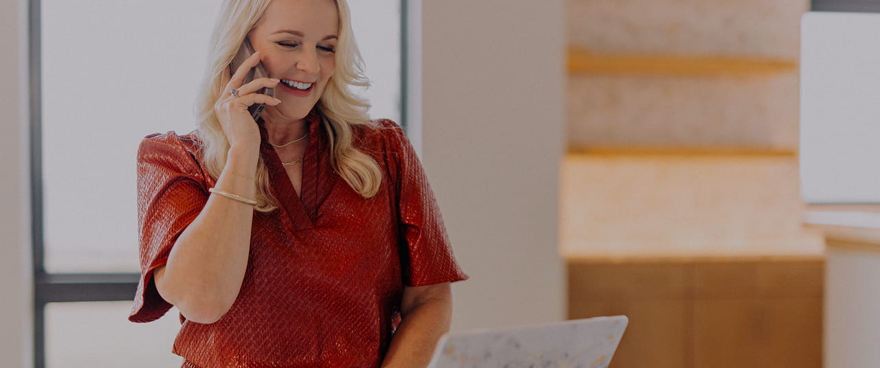 Smiling woman in a red blouse talking on a mobile phone while looking at a laptop.