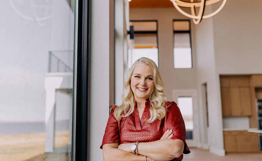Smiling woman with long blonde hair and red top standing with arms crossed by a window in a modern, well-lit interior.