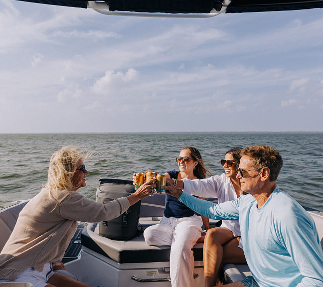 Four people sitting on a boat clinking cans in a toast on a sunny day at sea.