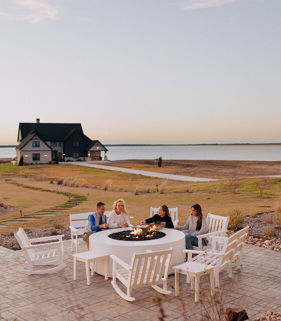 Four people sitting around a white outdoor fire pit with rocking chairs on a patio near a lake at dusk, with a large house in the background.