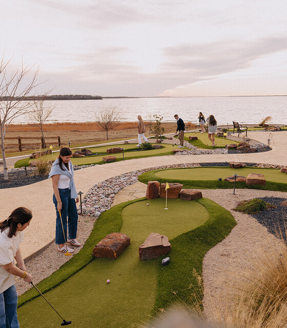 Group of people playing mini golf on a course near a lake during sunset.