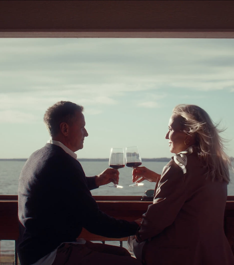 A man and woman sitting by a window overlooking water, clinking glasses of red wine during daytime.
