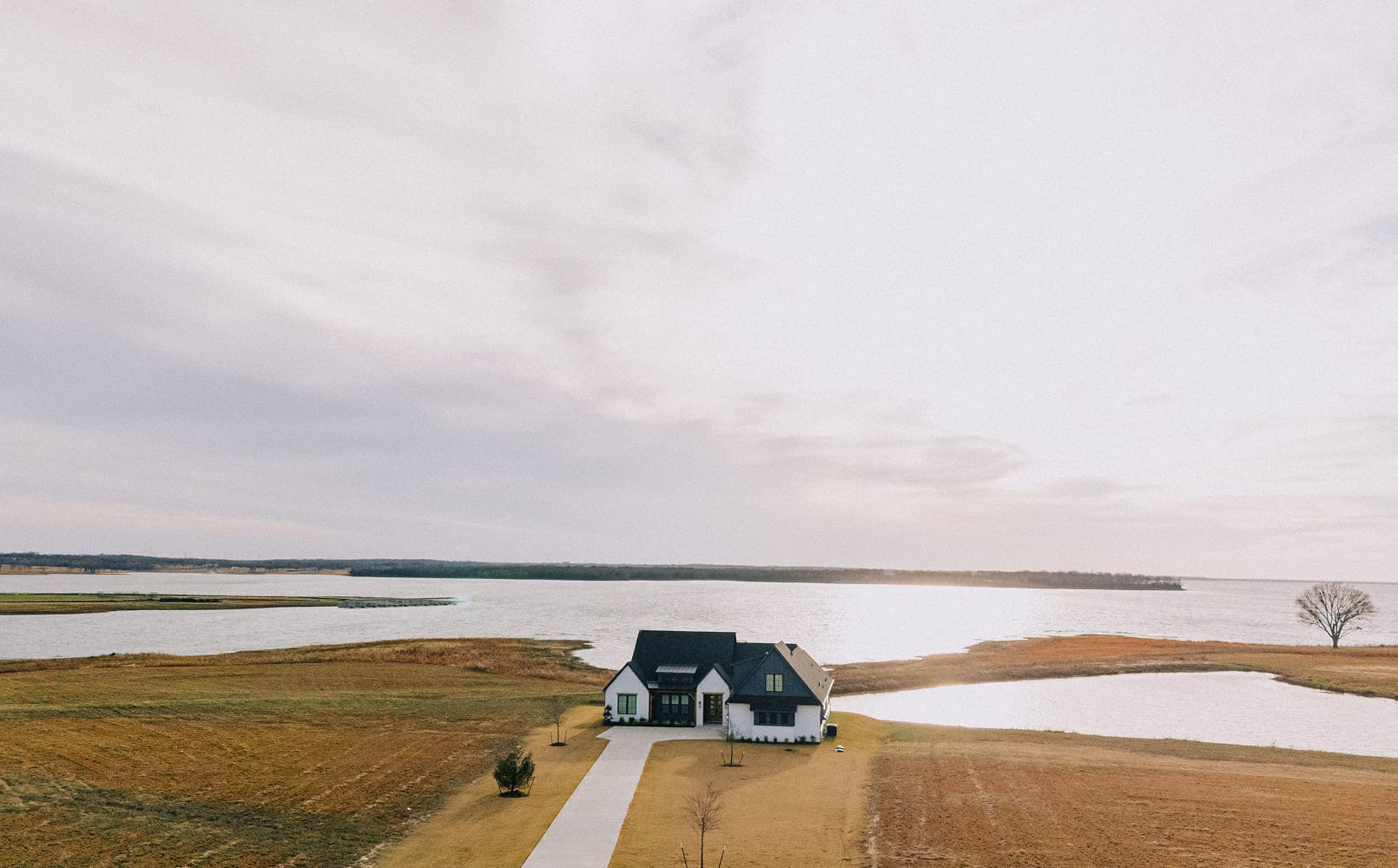 A modern white house with a dark roof situated on a grassy plot near a large body of water under a cloudy sky.