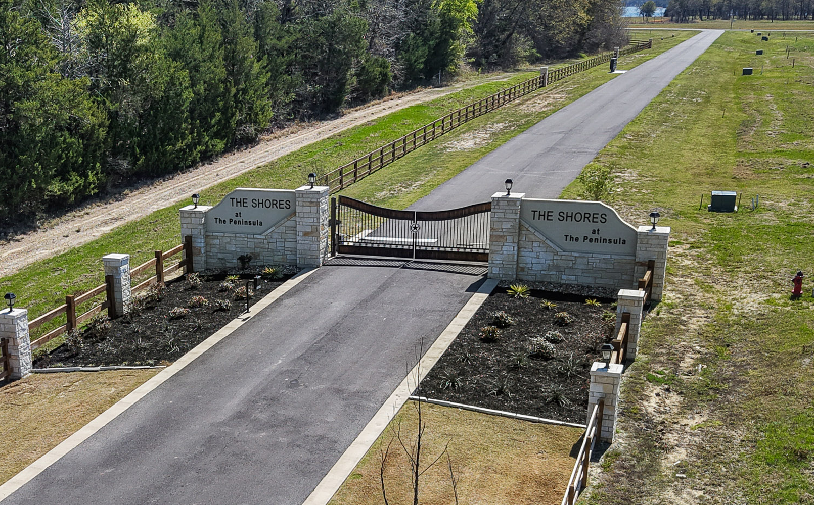 Gated entrance with stone pillars and signs reading 'THE SHORES at The Peninsula' flanking a paved road.