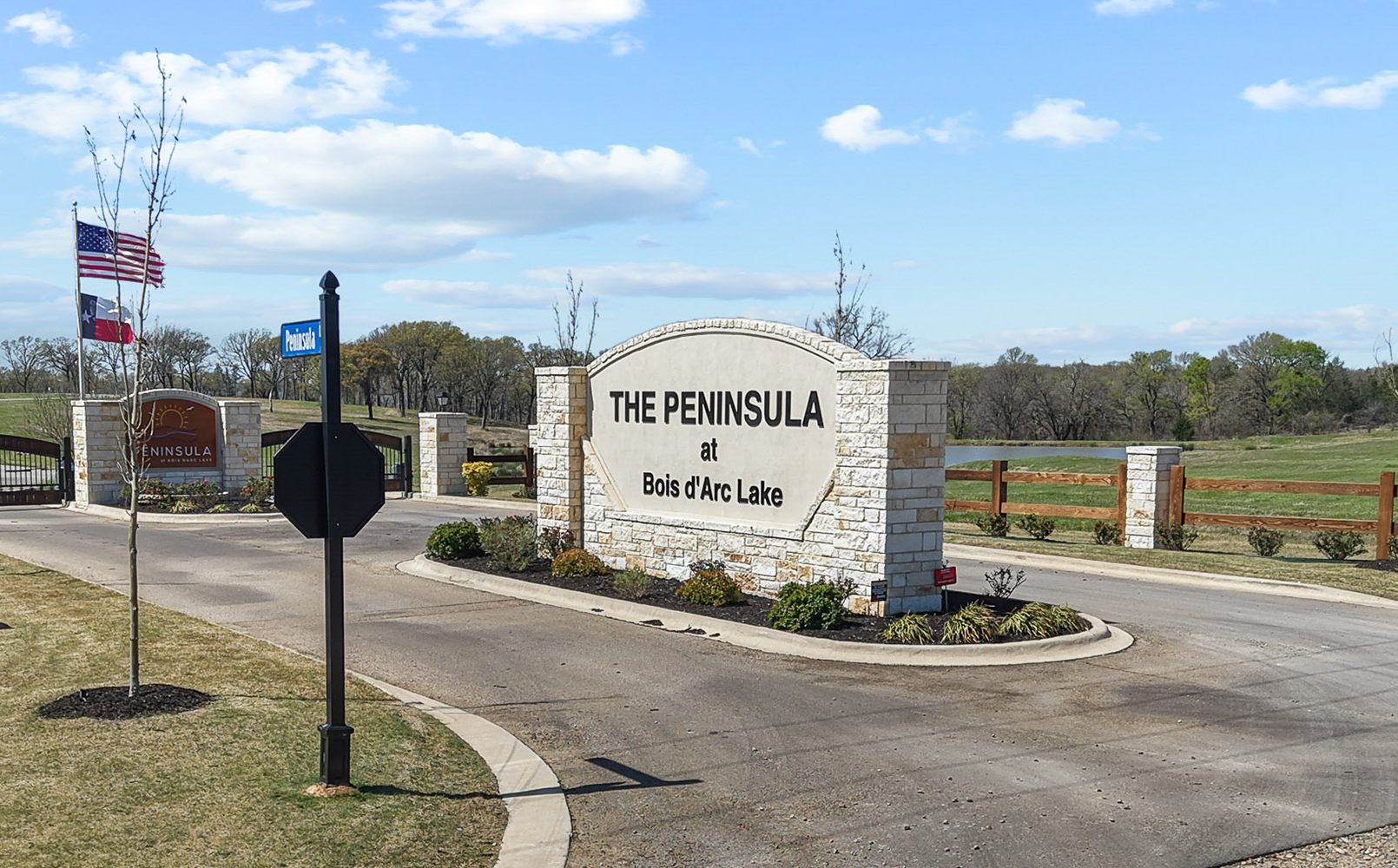 Entrance sign for The Peninsula at Bois d'Arc Lake with stone pillars, landscaped greenery, American and Texas flags, and a clear sky.