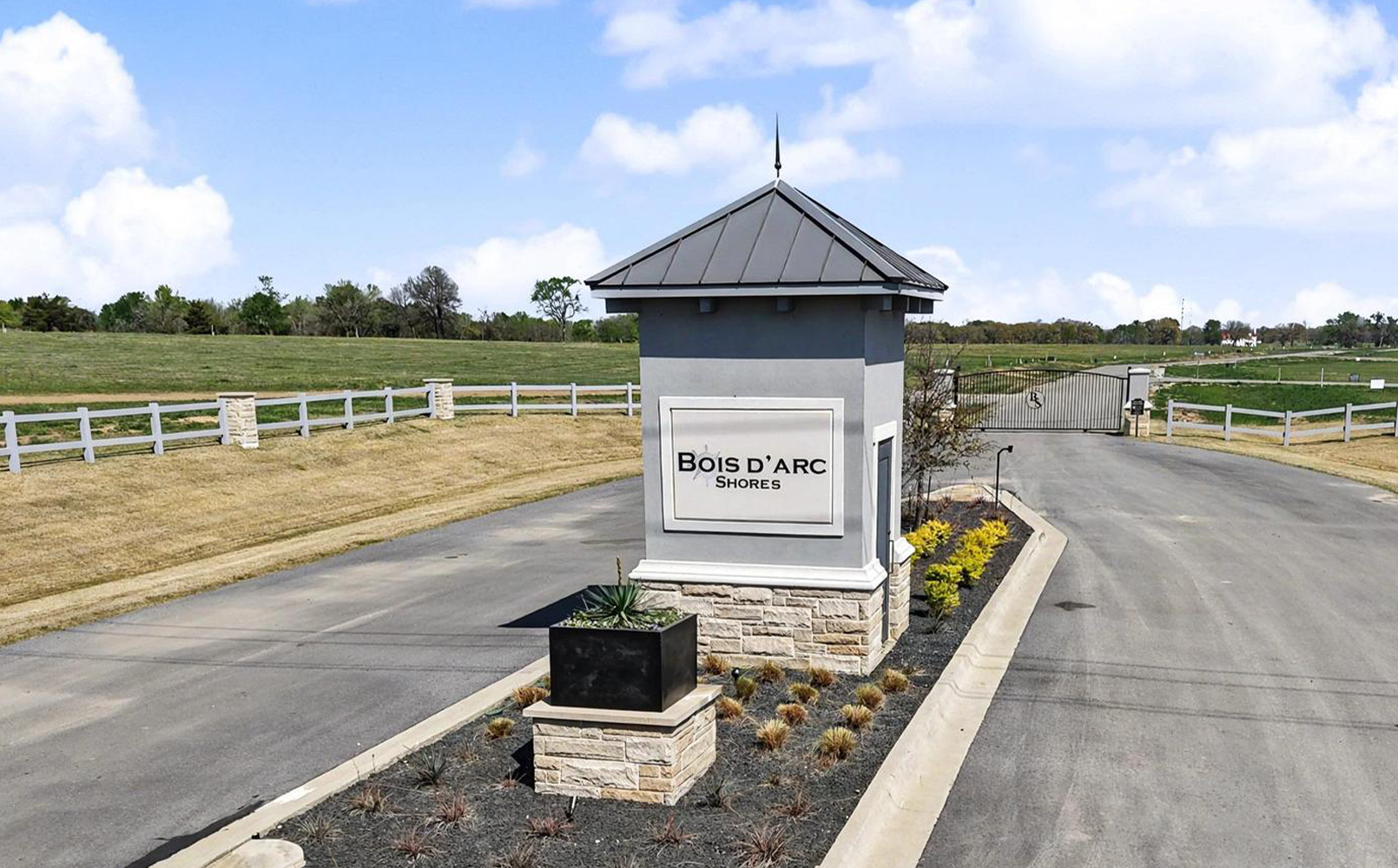 Entrance gate to Bois d'Arc Shores community with a gray stone sign and landscaped plants on a sunny day.