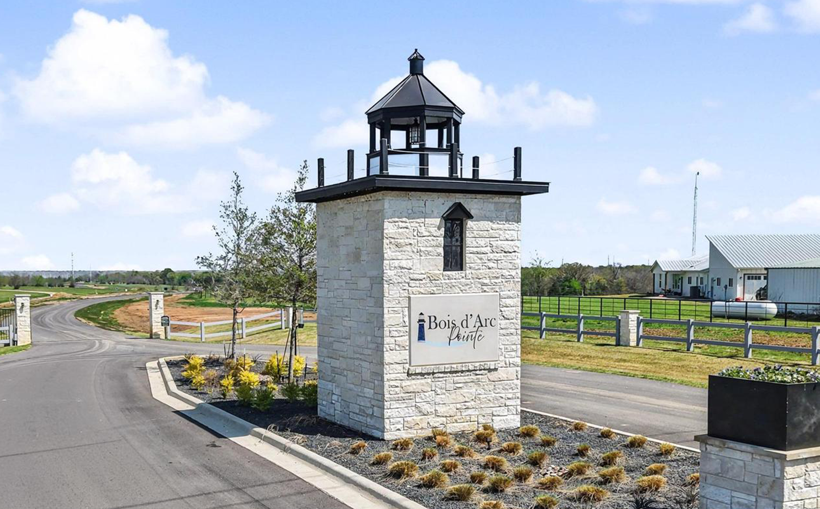 Stone entrance monument with black lantern top and Bois d'Arc Pointe sign beside a paved road and green fields under a blue sky.