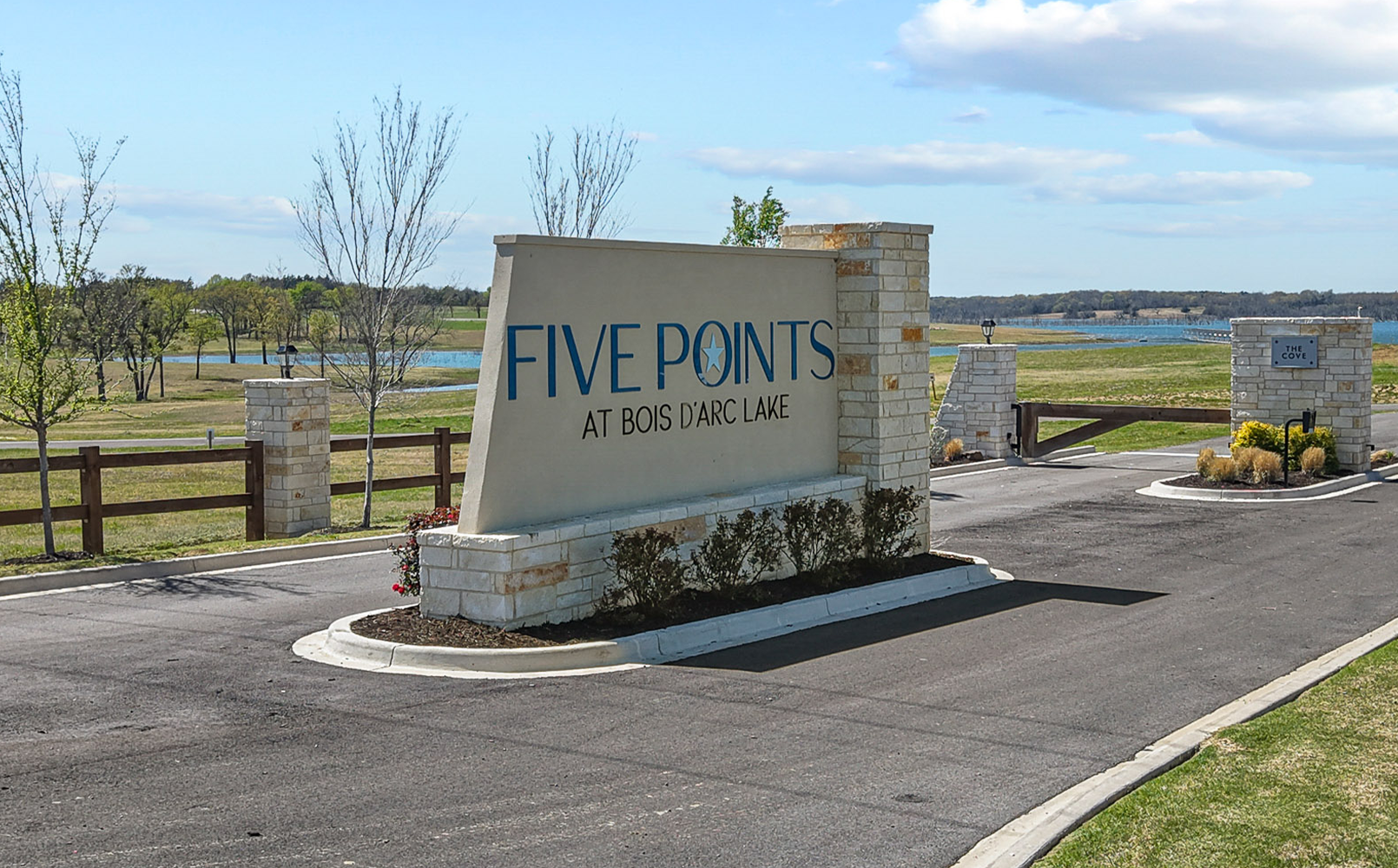 Entrance sign reading 'FIVE POINTS AT BOIS D'ARC LAKE' at a gated community road with a lake and trees in the background.