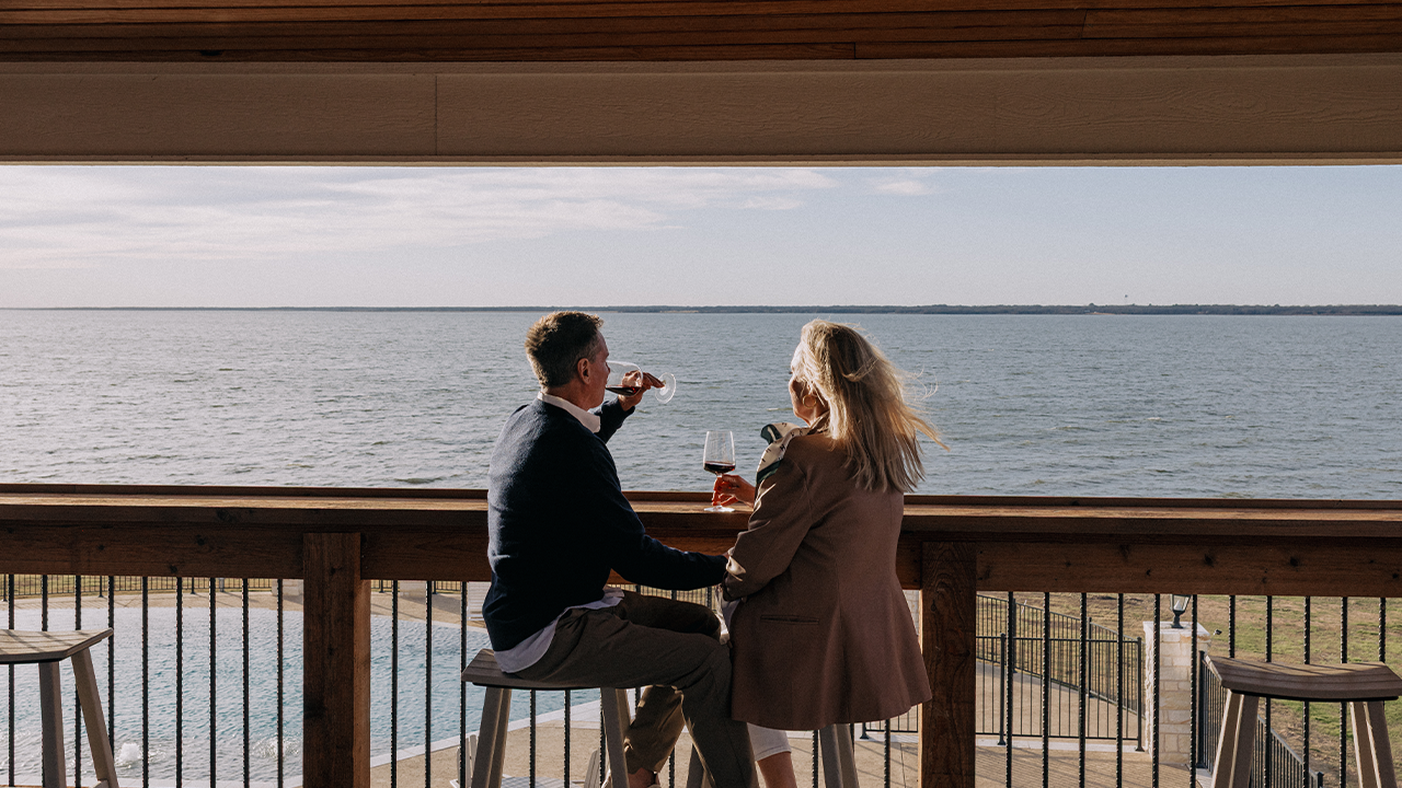 Man and woman sitting on bar stools, drinking red wine and overlooking a large body of water from a wooden balcony.