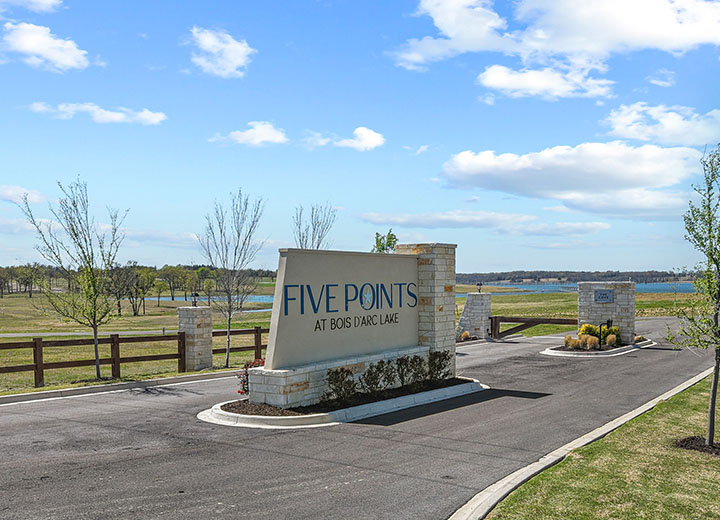 Entrance sign reading 'Five Points at Bois d'Arc Lake' on a stone and concrete structure beside a paved road with a lake and trees in the background under a partly cloudy sky.