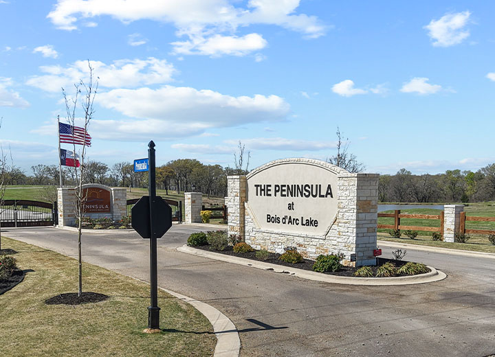 Entrance sign for The Peninsula at Bois d'Arc Lake with stone pillars, an American flag, a Texas flag, and a blue street sign under a partly cloudy sky.