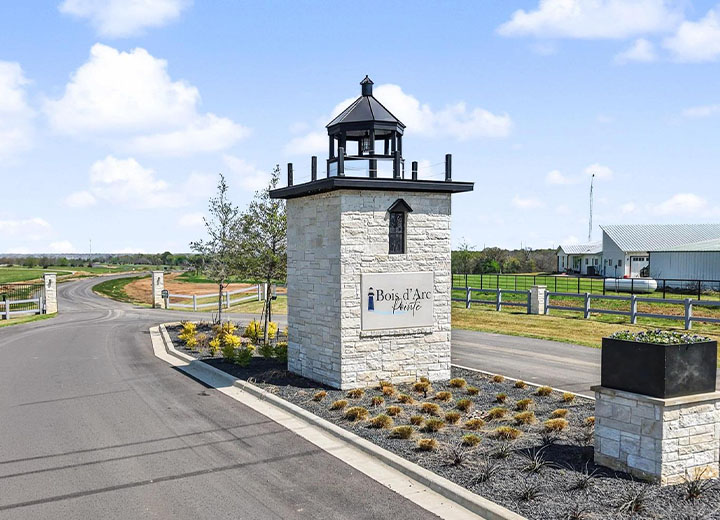 Stone entrance monument with a lantern-style top and sign reading 'Bois d'Arc Pointe' beside a curving paved road and landscaped greenery under a partly cloudy sky.