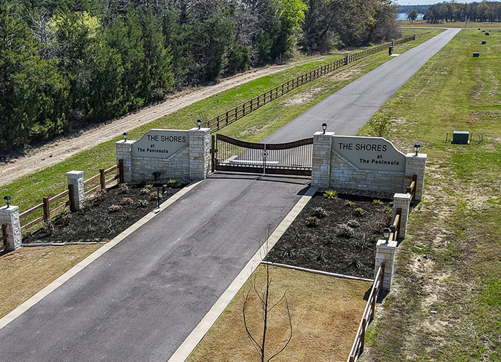 Gated entrance to The Shores at The Peninsula community with stone pillars, black metal gate, and a long paved road surrounded by grass and trees.