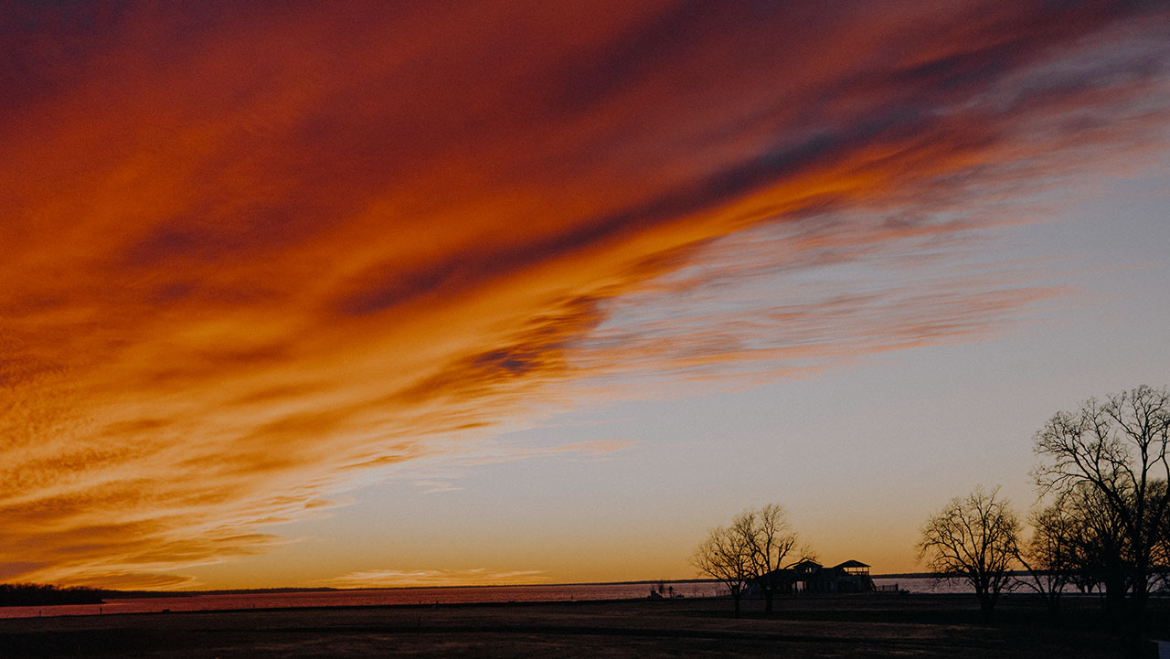 Silhouette of trees and a house under a dramatic orange and purple sunset sky over a flat landscape.