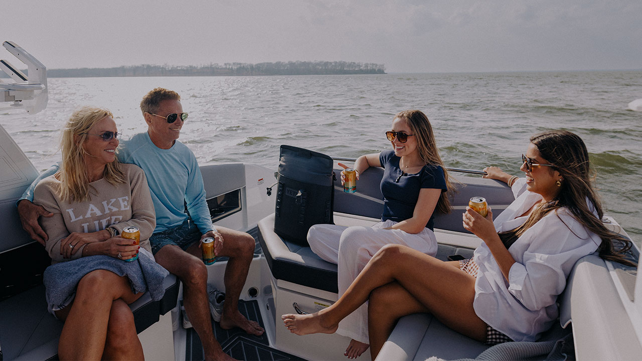 Four adults wearing sunglasses sitting and relaxing on a boat, holding canned drinks with a lake and distant tree line in the background.