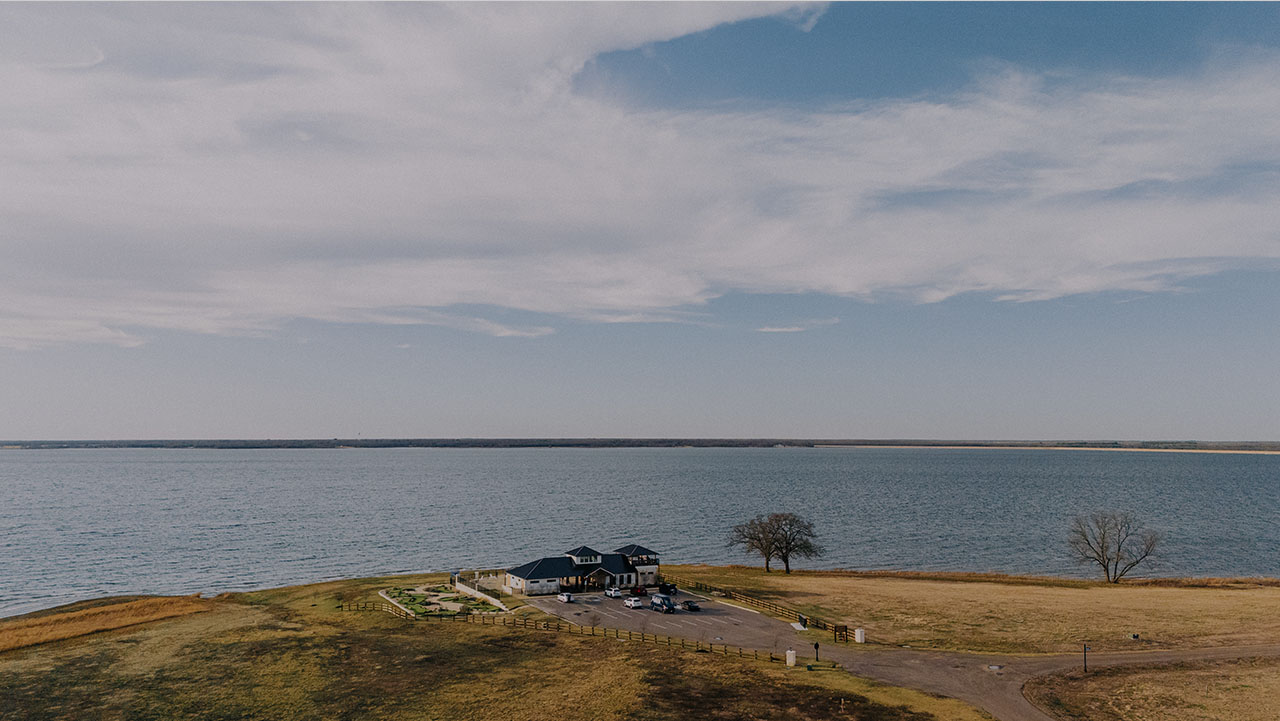 A small building with parked cars beside a large body of water under a partly cloudy sky.