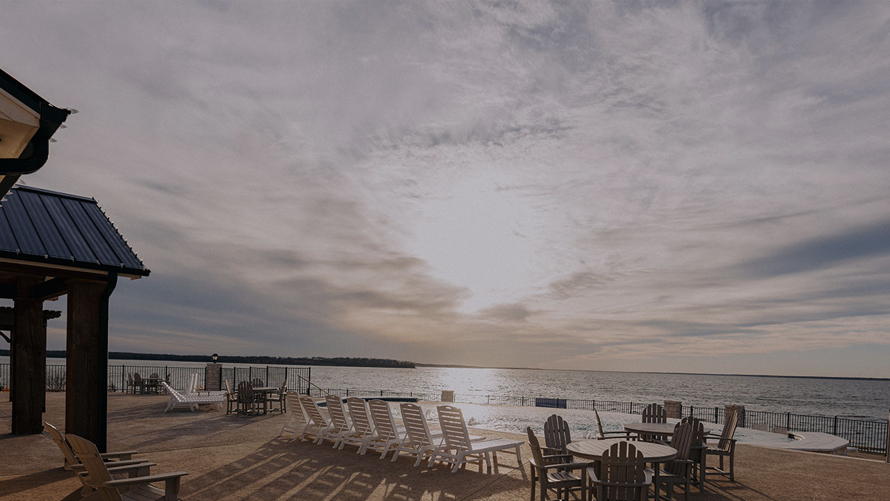 Outdoor pool area with lounge chairs and round tables facing a large body of water under a cloudy sky at sunset.