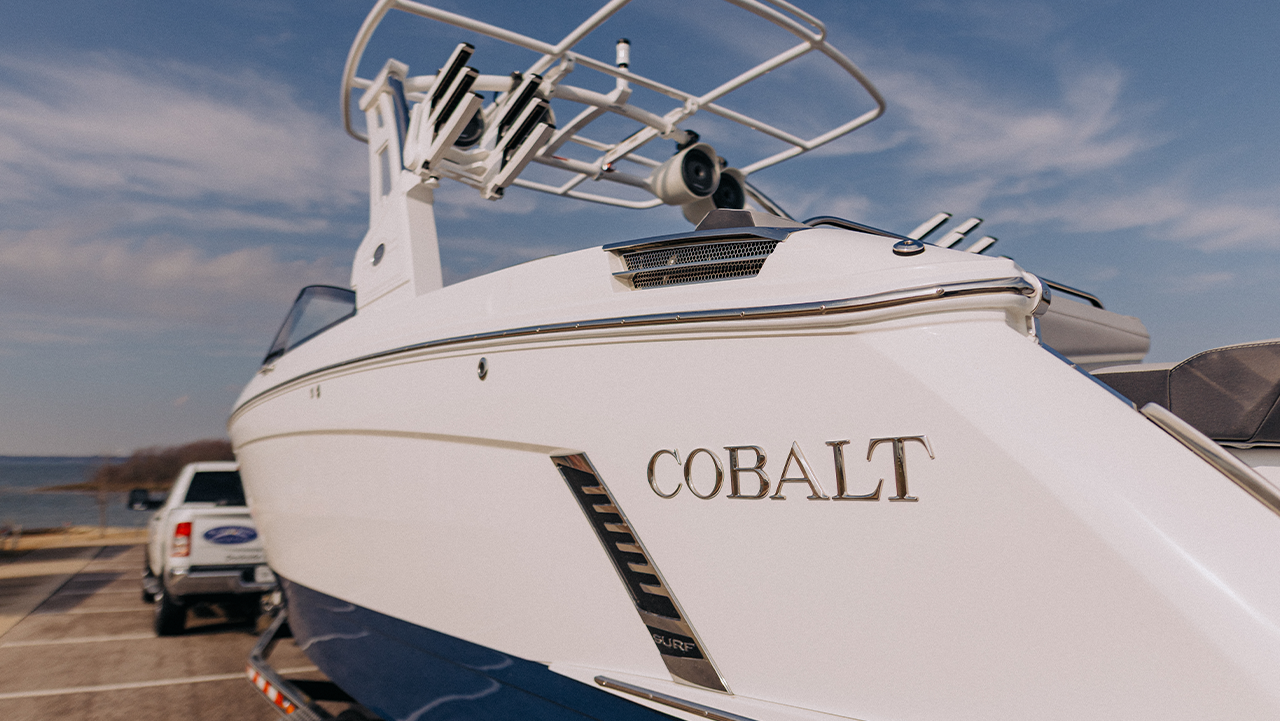 Close-up of a white and blue Cobalt motorboat on a trailer with a truck in the background under a partly cloudy sky.