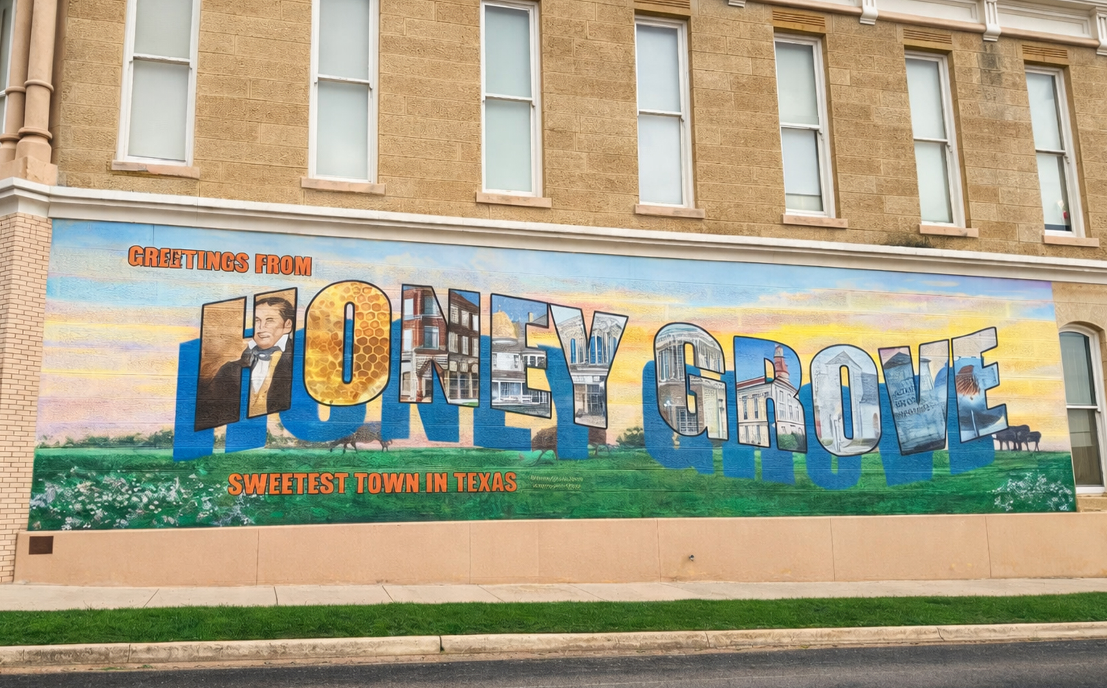 A mural on a brick wall reading 'Greetings from Honey Grove, Sweetest Town in Texas' with each letter containing images of local scenes and icons.