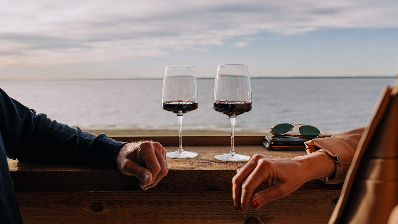 Two hands resting on a wooden surface with two glasses of red wine and a pair of sunglasses by the water.