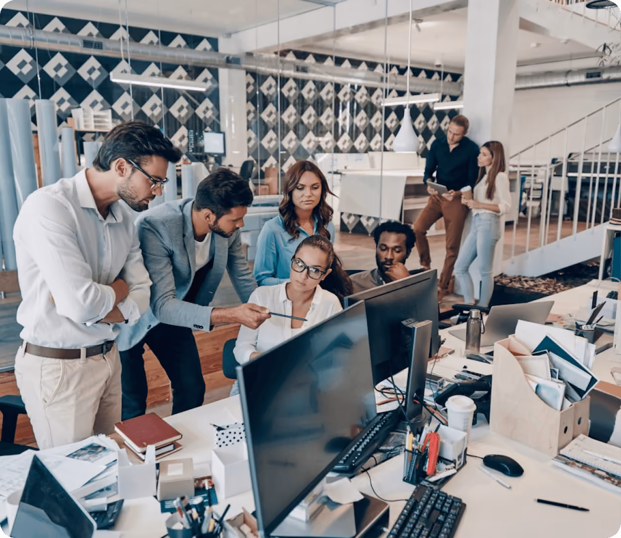 Groupe de six collègues discutant autour d'ordinateurs dans un bureau moderne et lumineux.