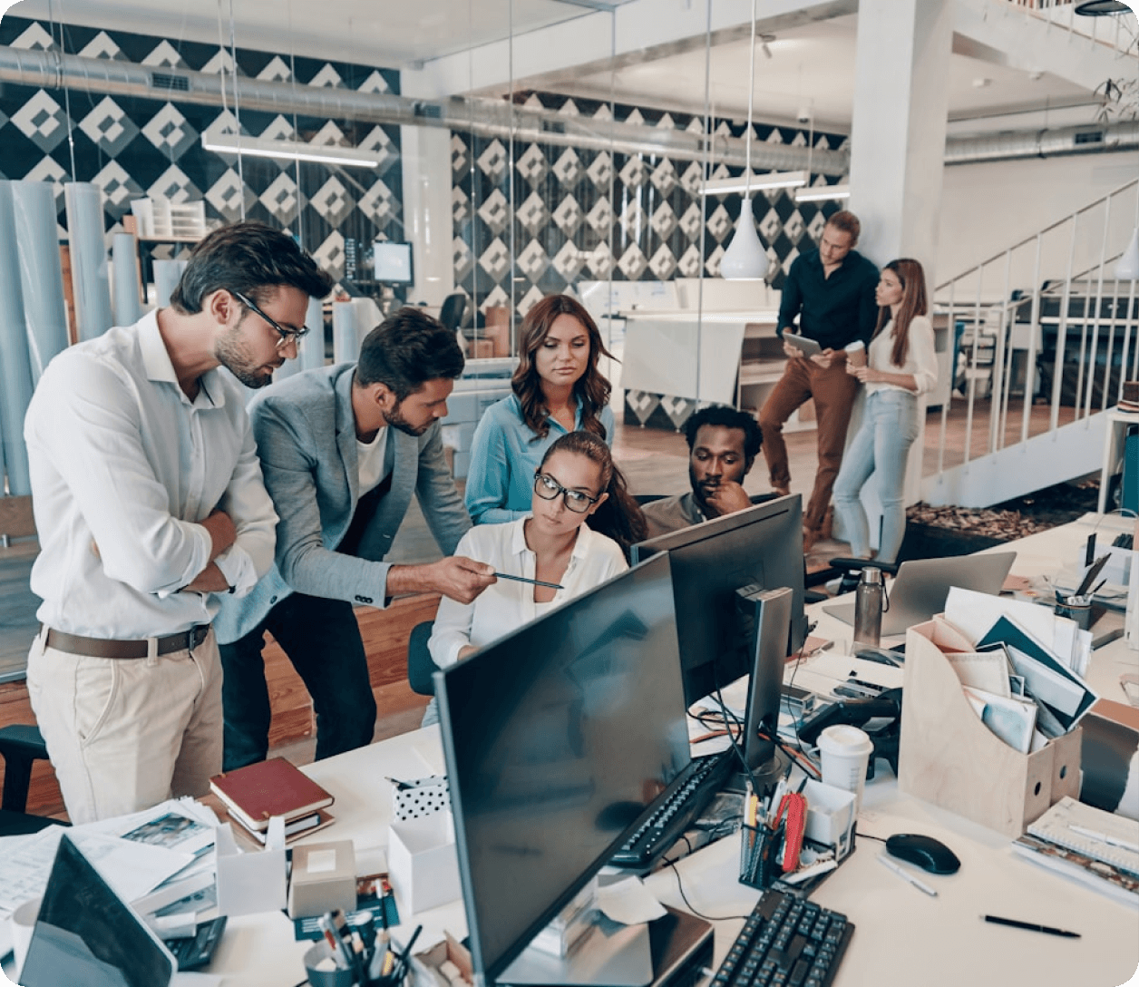 Groupe de six collègues discutant autour d'ordinateurs dans un bureau moderne et lumineux.