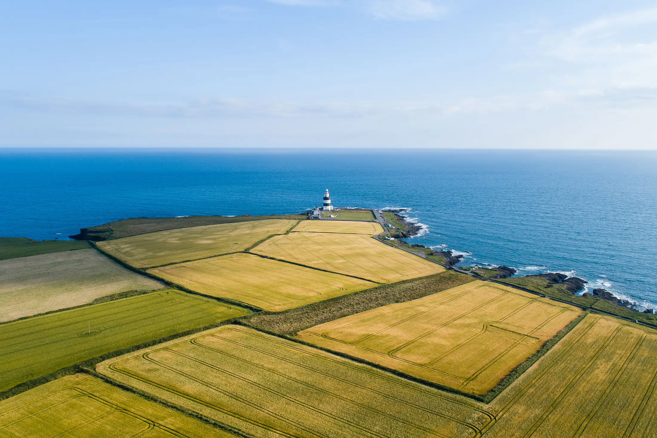 Aerial view of patchwork farmland leading to a lighthouse on a narrow coastal promontory with the ocean in the background.
