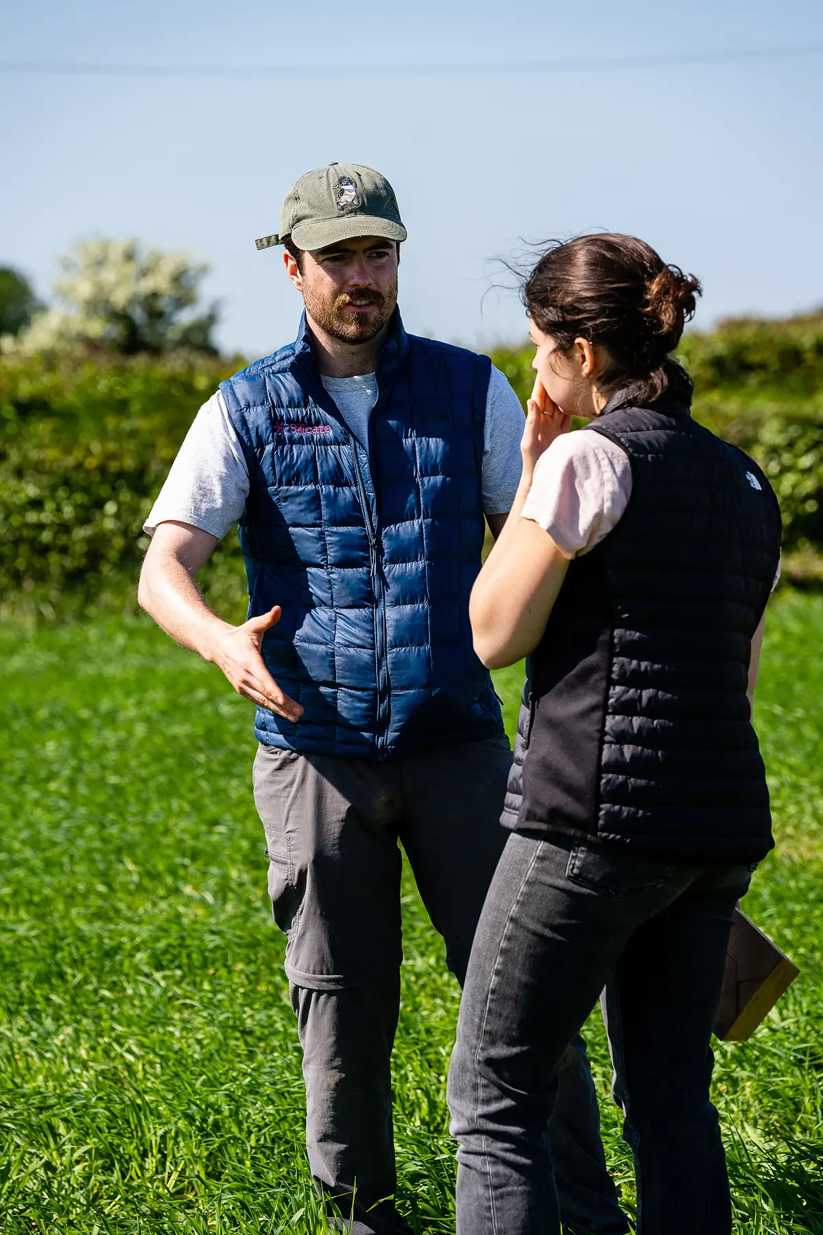 Man and woman in outdoor field having a conversation, both wearing quilted vests.