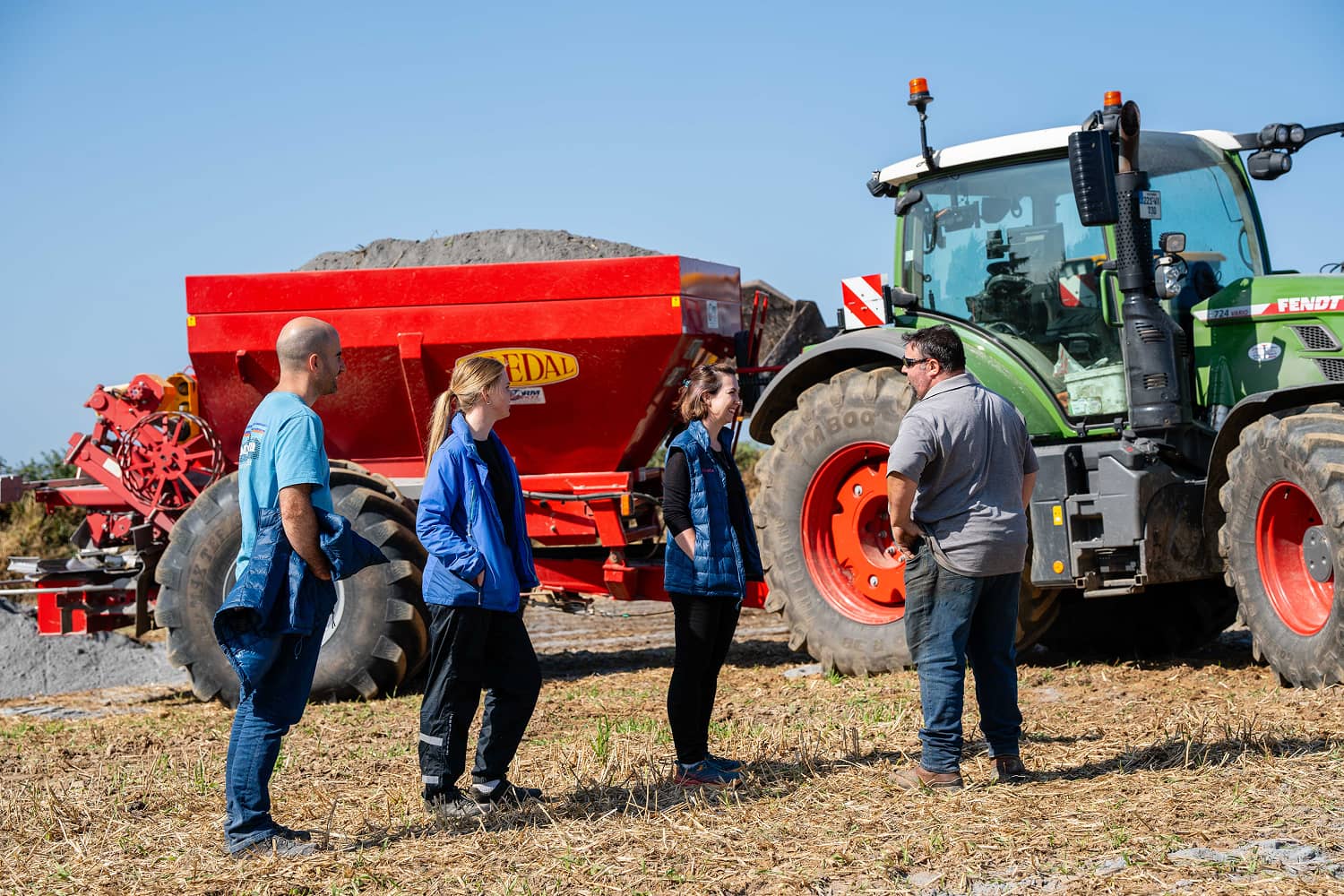 Four people standing and talking near a green tractor with a red farming trailer filled with soil under a clear blue sky.
