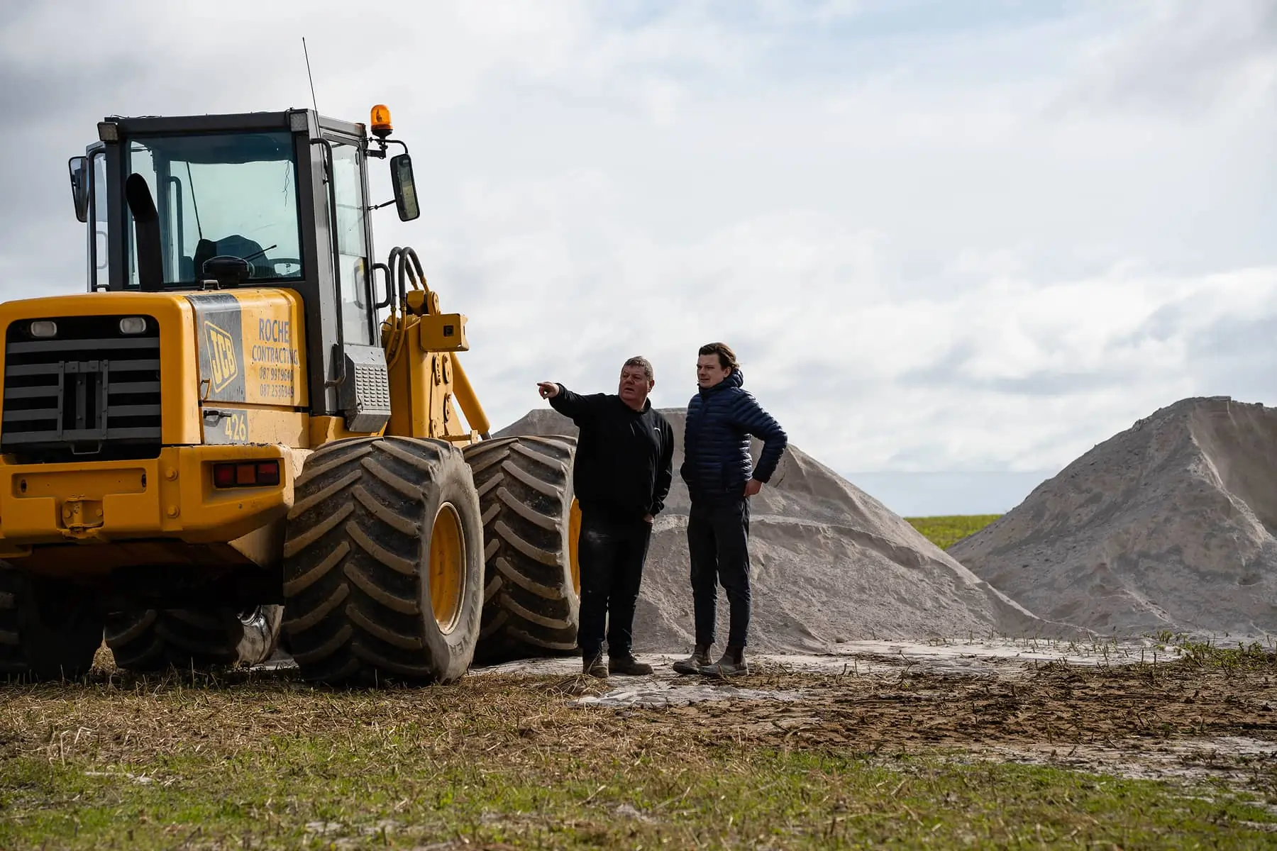 Two men standing on a field next to a large yellow construction vehicle with piles of sand in the background.