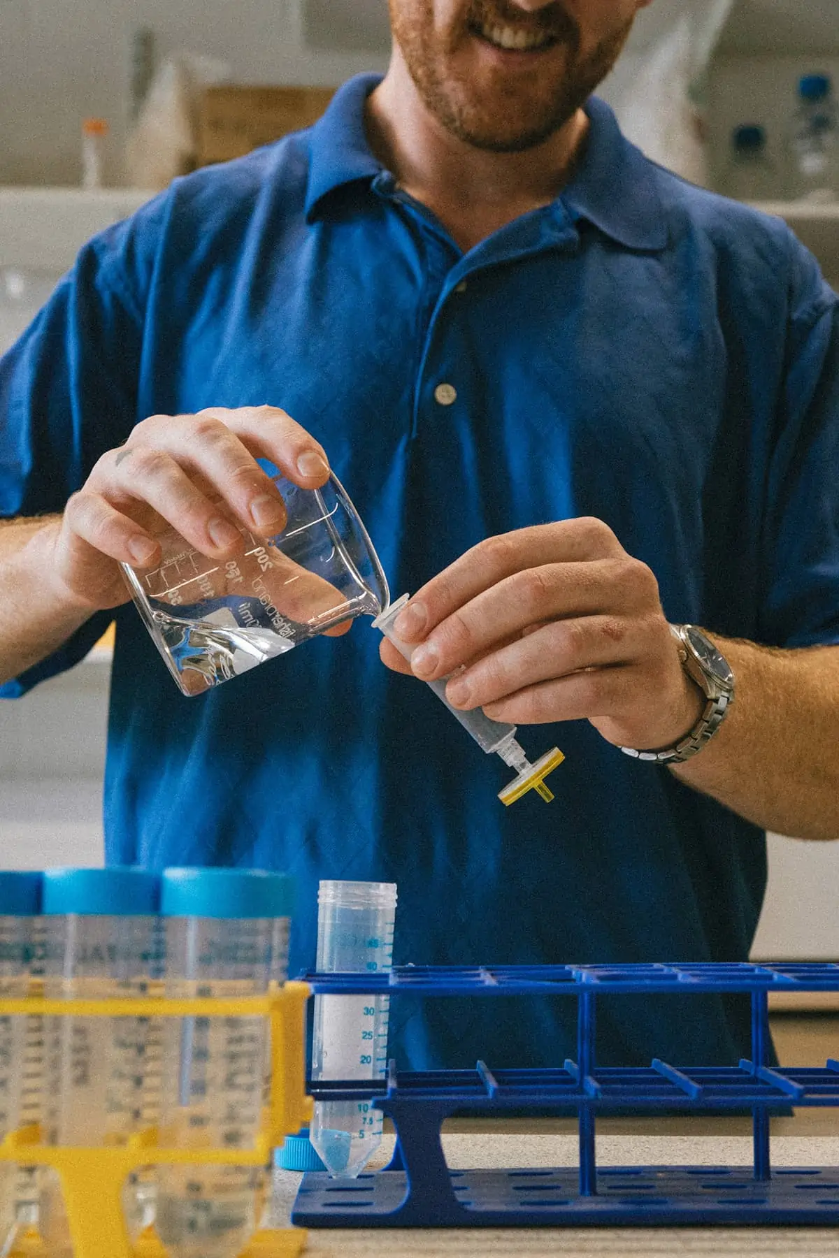 Person in blue shirt pouring liquid from a beaker into a syringe in a laboratory setting.