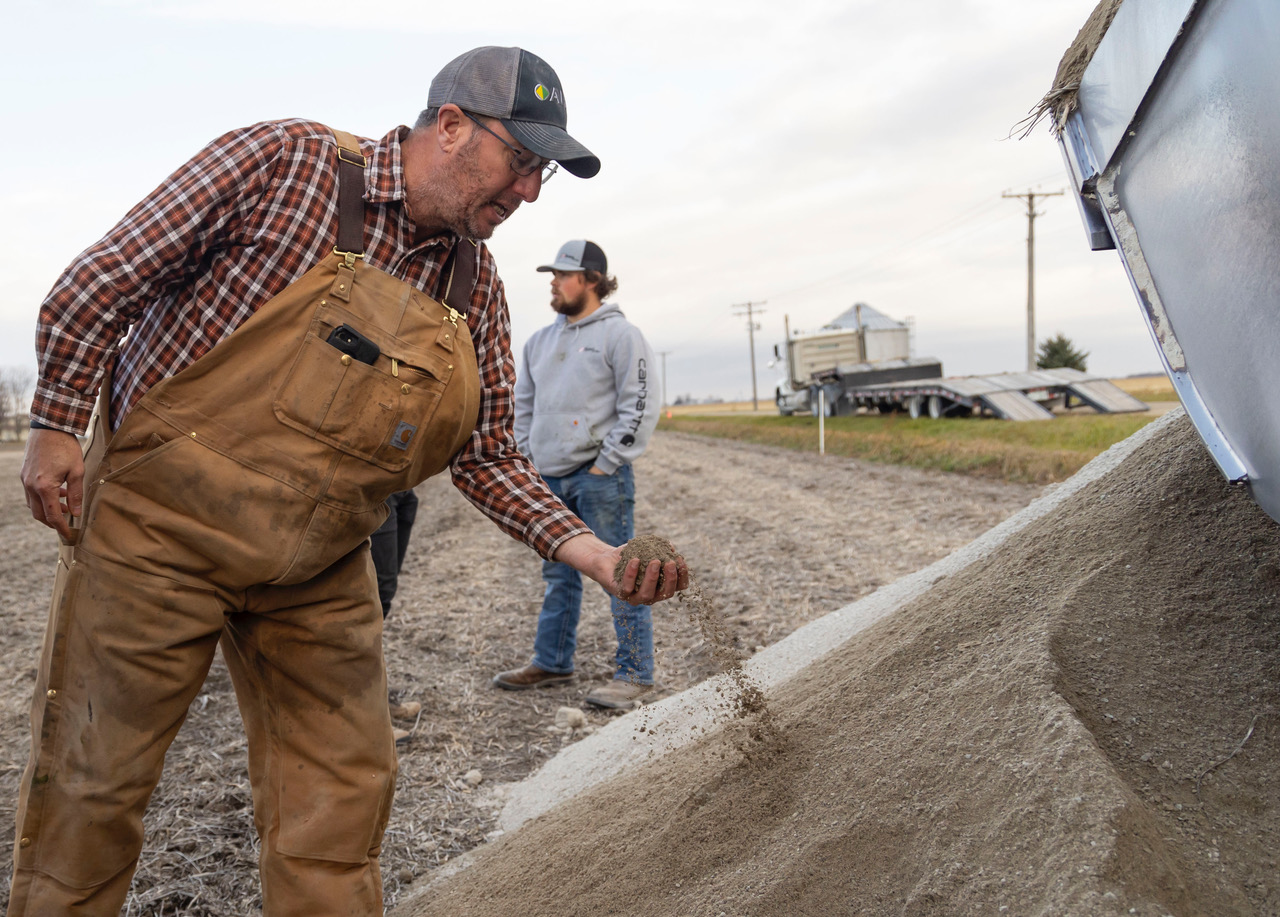 Man in plaid shirt and brown overalls examining and sifting sandy soil from a truck while another man stands in the background on a farm.