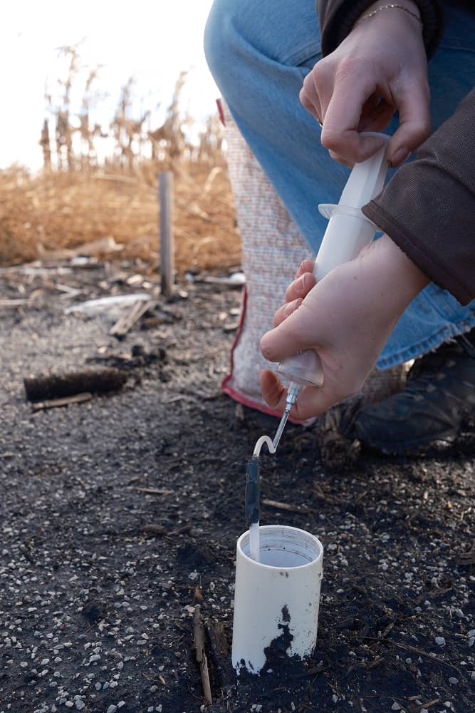 Person using a syringe to collect a soil sample from a white PVC pipe inserted in the ground.