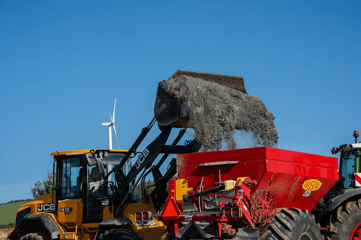 Yellow JCB loader dumping soil into a large red Bredal agricultural spreader attached to a tractor under a clear blue sky.