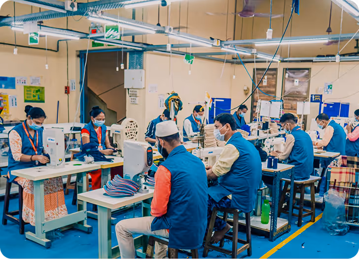Workers wearing masks and blue vests sewing garments on machines in a garment factory.